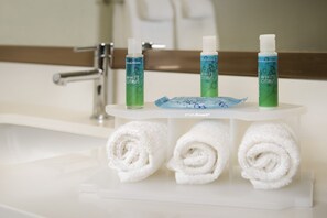 Un baño con un lavamanos blanco, tres botellas de gel de ducha White Citrus y tres toallas blancas enrolladas sobre una estantería blanca.