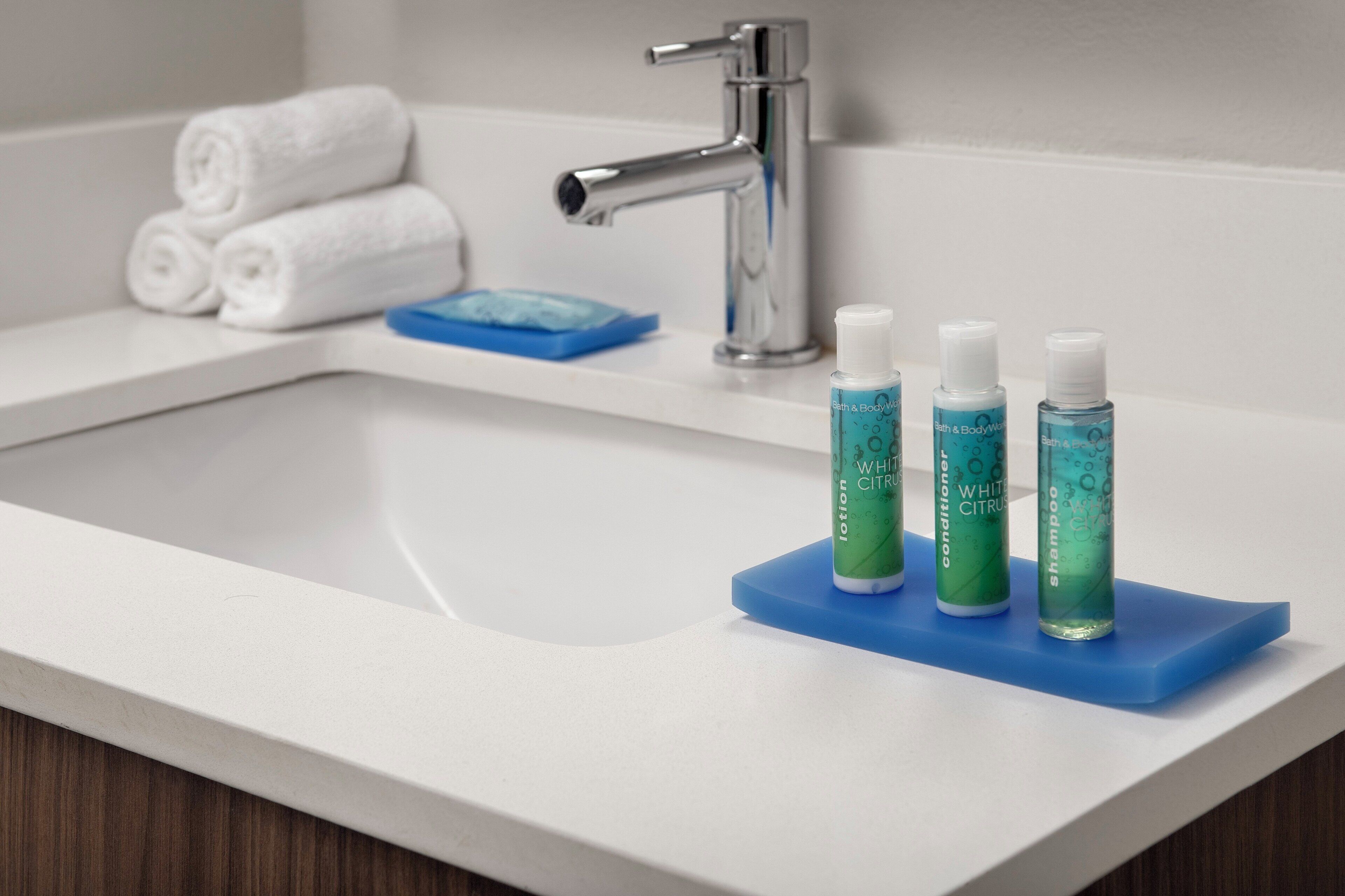 Uma pia de banheiro com três frascos de produtos de higiene pessoal em um porta-sabonete azul, uma torneira prateada e toalhas brancas dobradas em uma prateleira.