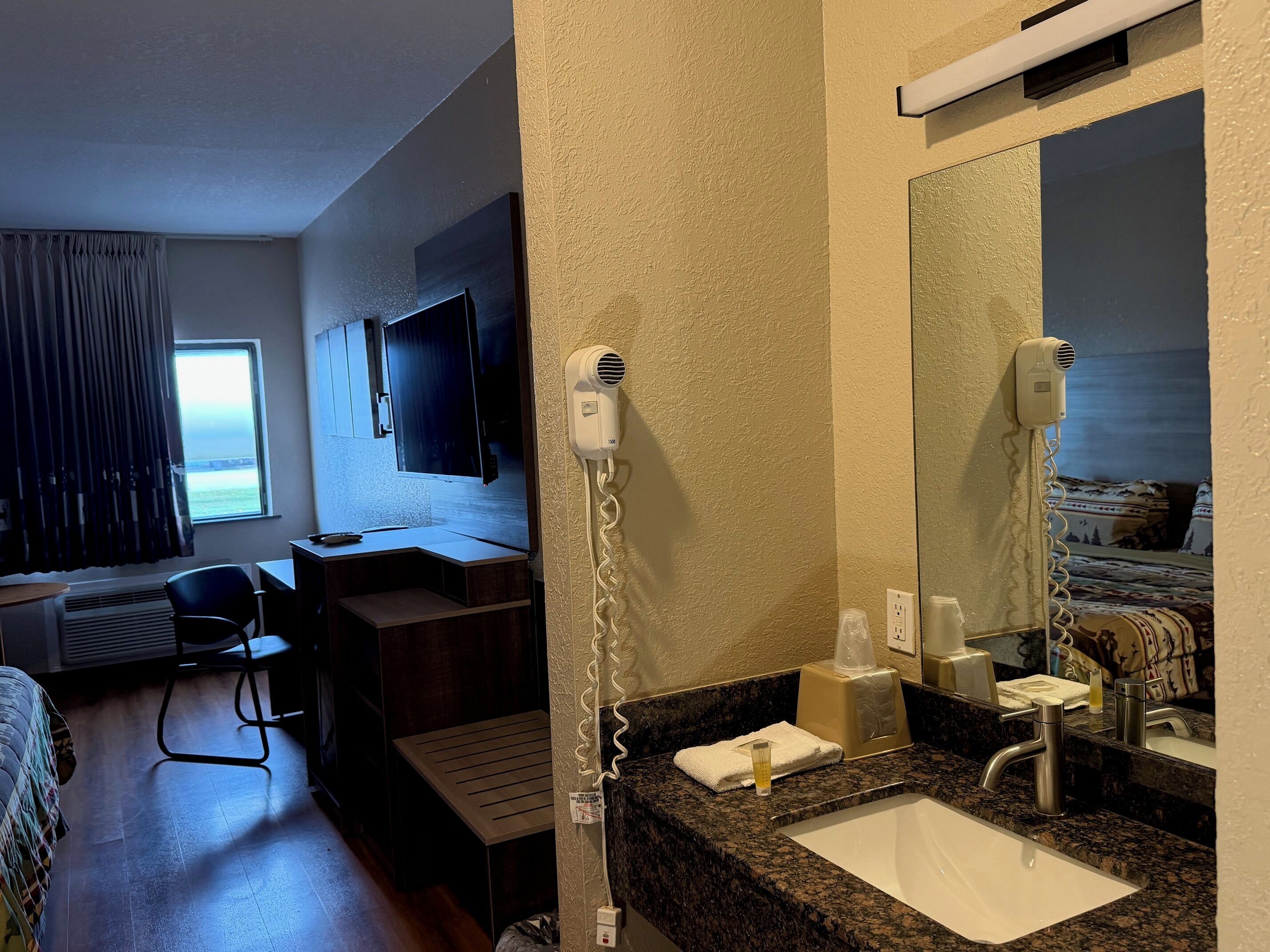 Une chambre d’hôtel équipée d’un lit, d’un bureau, d’une chaise, d’une télévision, d’un sèche-cheveux, d’un lavabo et d’une fenêtre avec des rideaux.