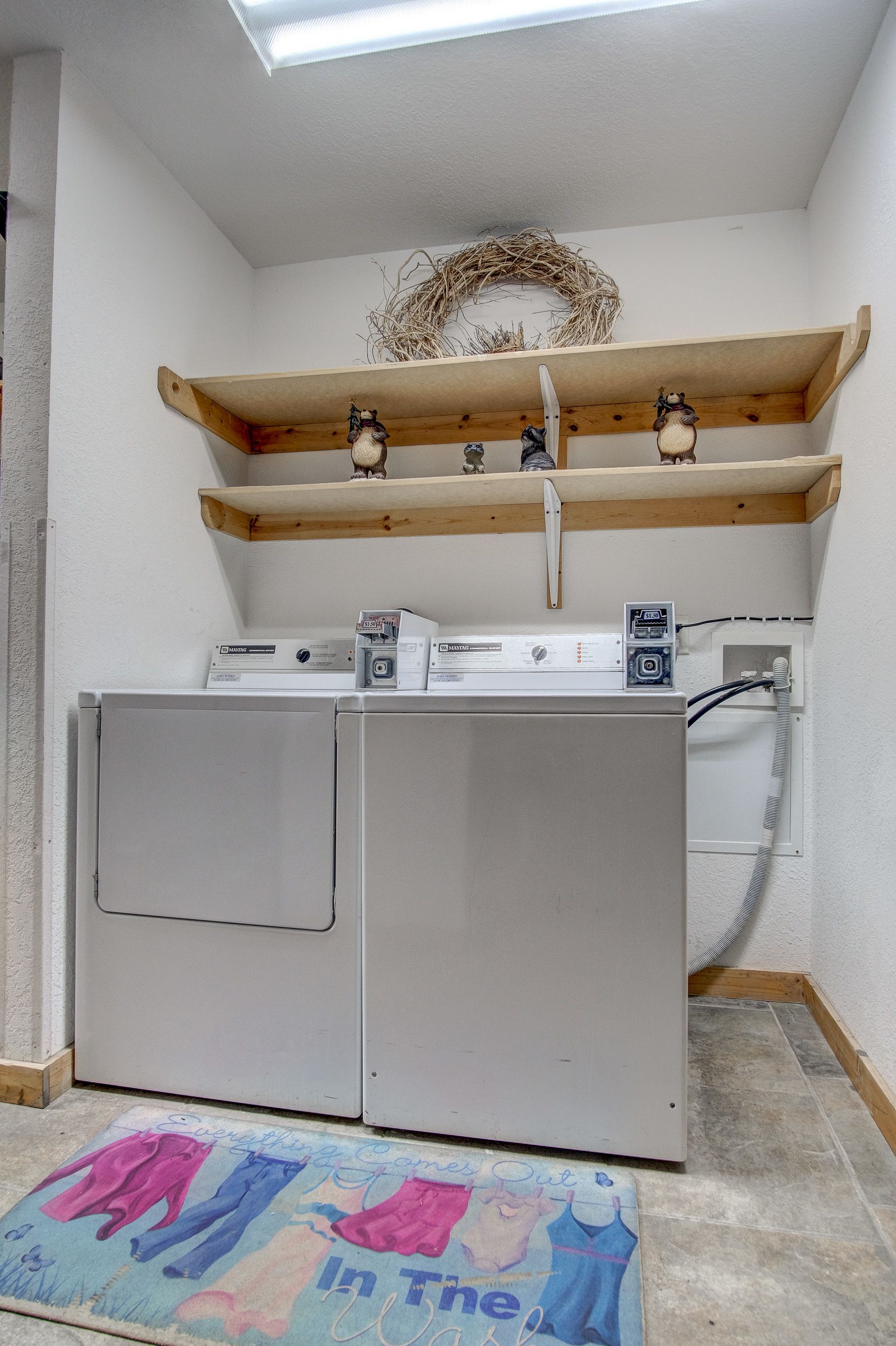 A laundry room with two front-loading washing machines, wooden shelves above, and a door mat with a cartoon character.