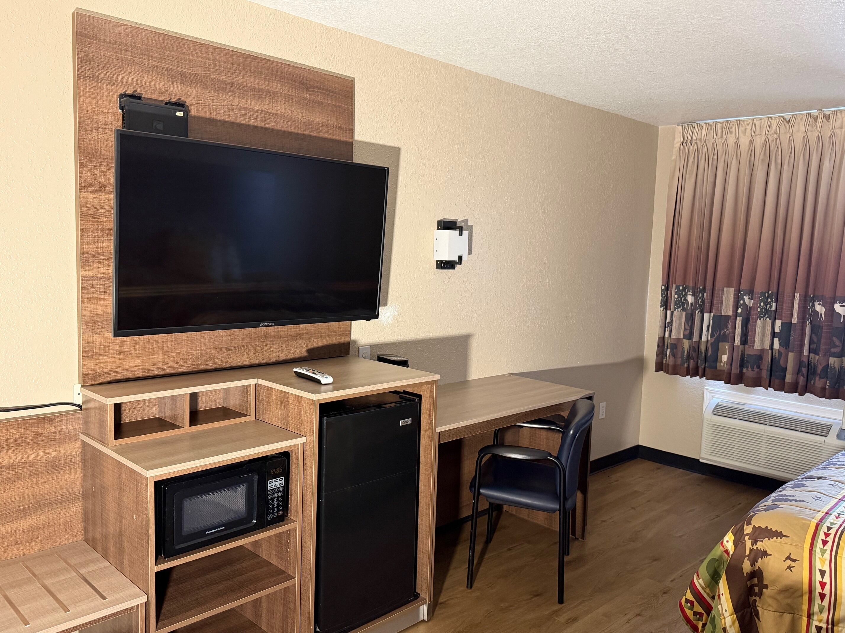 A hotel room with a flat-screen TV mounted on a wooden wall, a small refrigerator, a microwave, a desk with a chair, and a bed with a patterned bedspread.