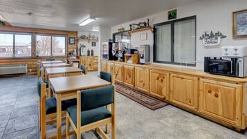 Un comedor con mesas y sillas de madera, una encimera de cocina con electrodomésticos y ventanas amplias.
