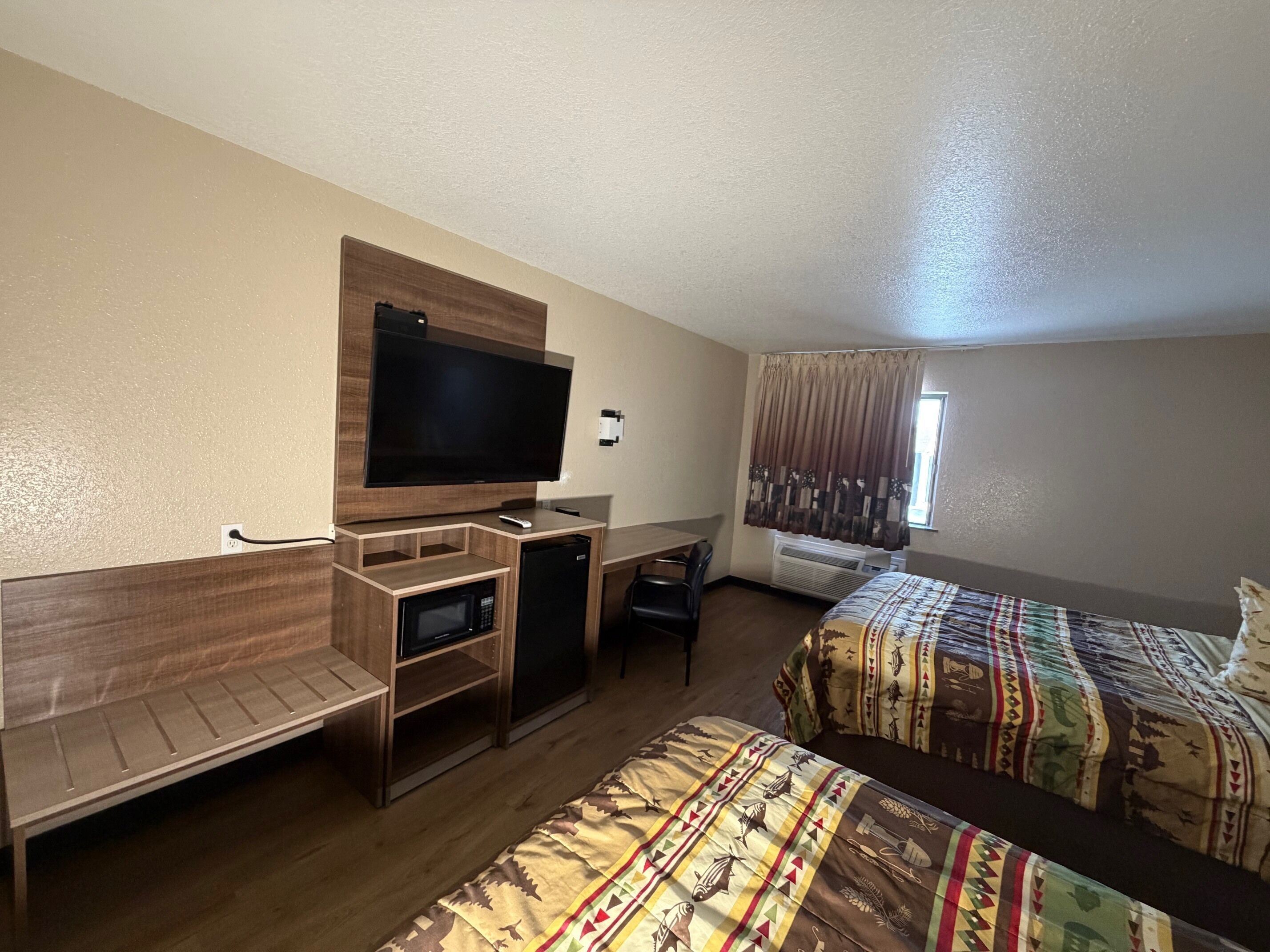 Une chambre d’hôtel équipée d’un téléviseur à écran plat, d’un bureau avec une chaise, d’un four à micro-ondes, d’un réfrigérateur, de deux lits avec une literie à motifs et d’une fenêtre avec des rideaux.