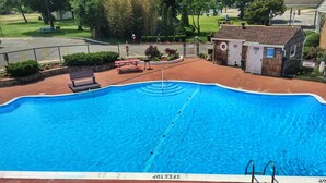 A swimming pool with a diving board, surrounded by a fenced patio area with benches and a small building.
