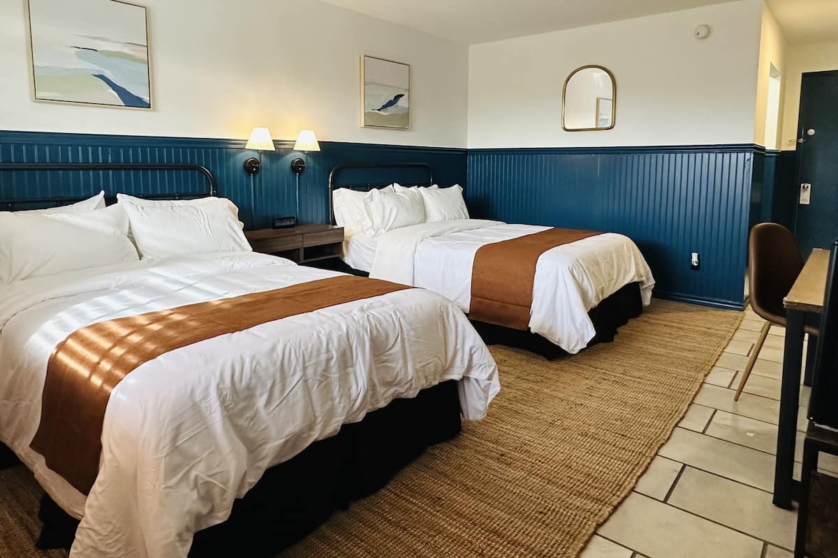 A hotel room with two beds, a small table, and a chair. There are wall-mounted lamps and a mirror above the beds.
