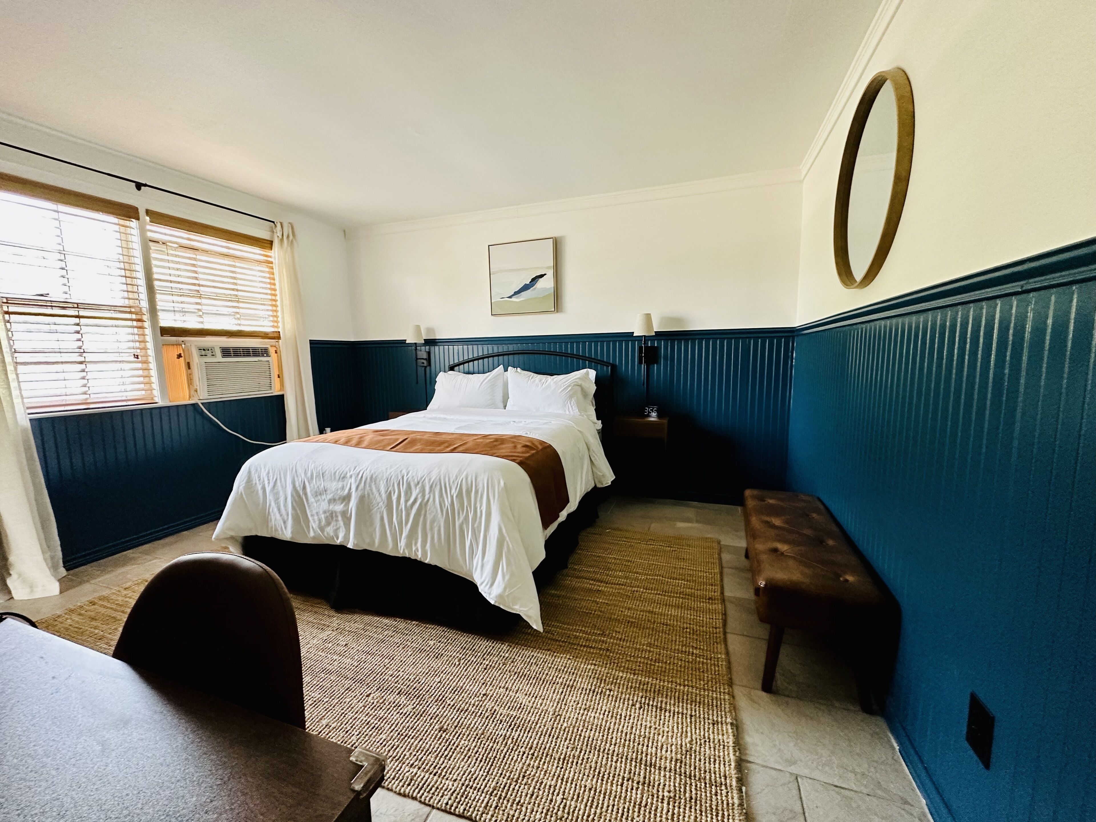 A hotel room with a bed, a desk, a bench, a window with blinds, and a wall-mounted lamp.