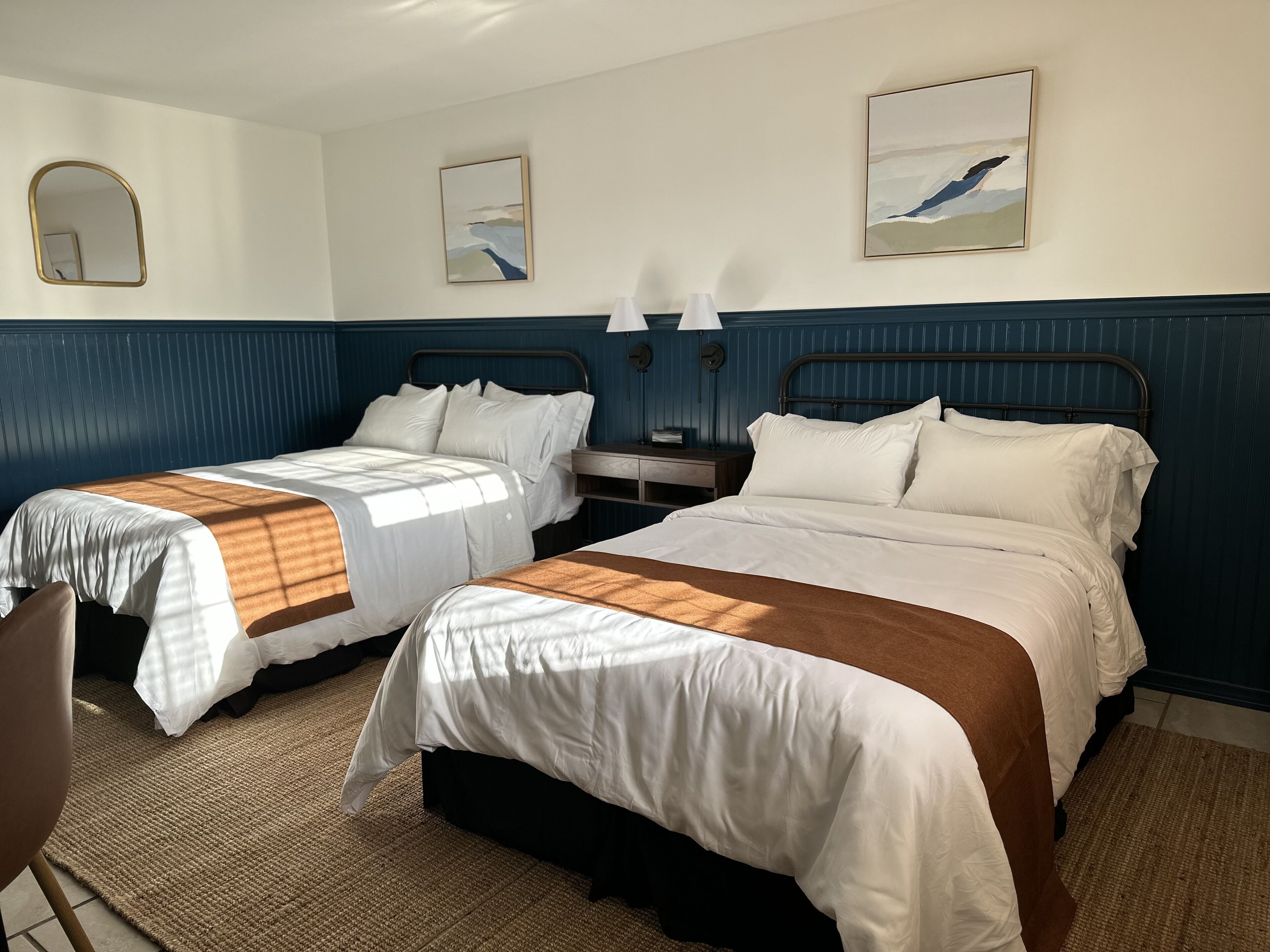 Quarto de hotel com duas camas, uma mesinha de cabeceira, uma cadeira e obras de arte nas paredes.