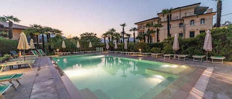 A swimming pool with lounge chairs and umbrellas, surrounded by greenery and a building with balconies.
