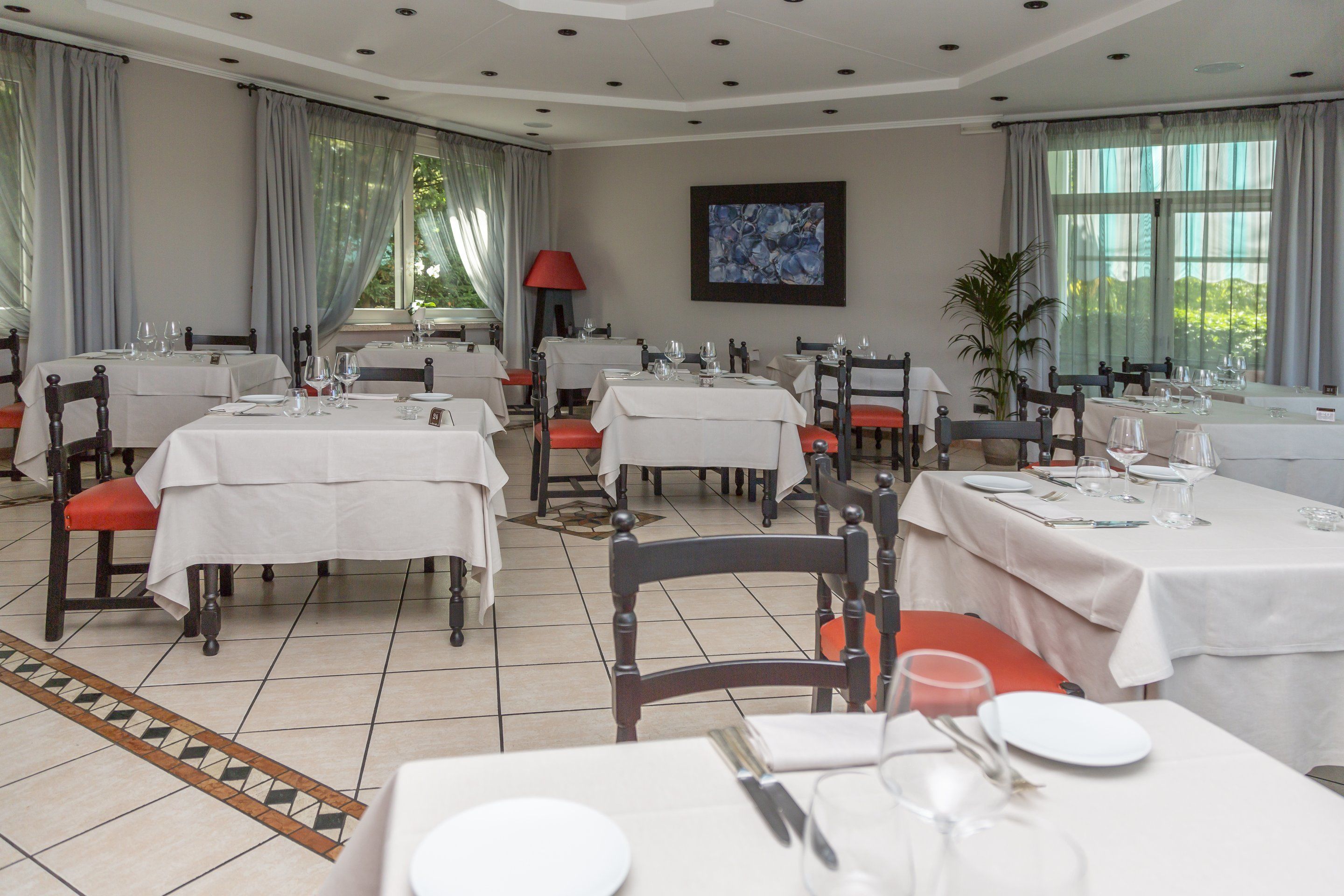 A restaurant with multiple tables set for dining, featuring a tiled floor and large windows with curtains.