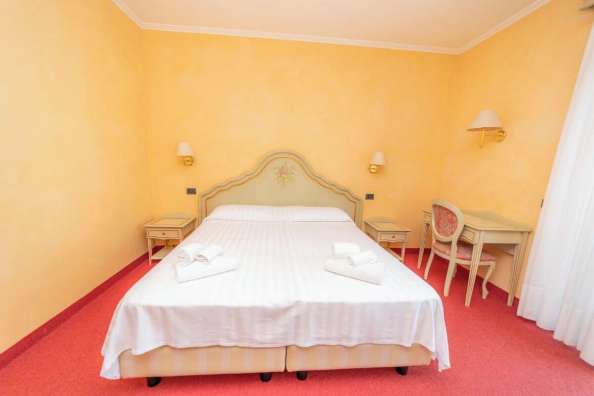 A hotel room with a double bed, two bedside tables, a desk with a chair, and two wall-mounted lamps.