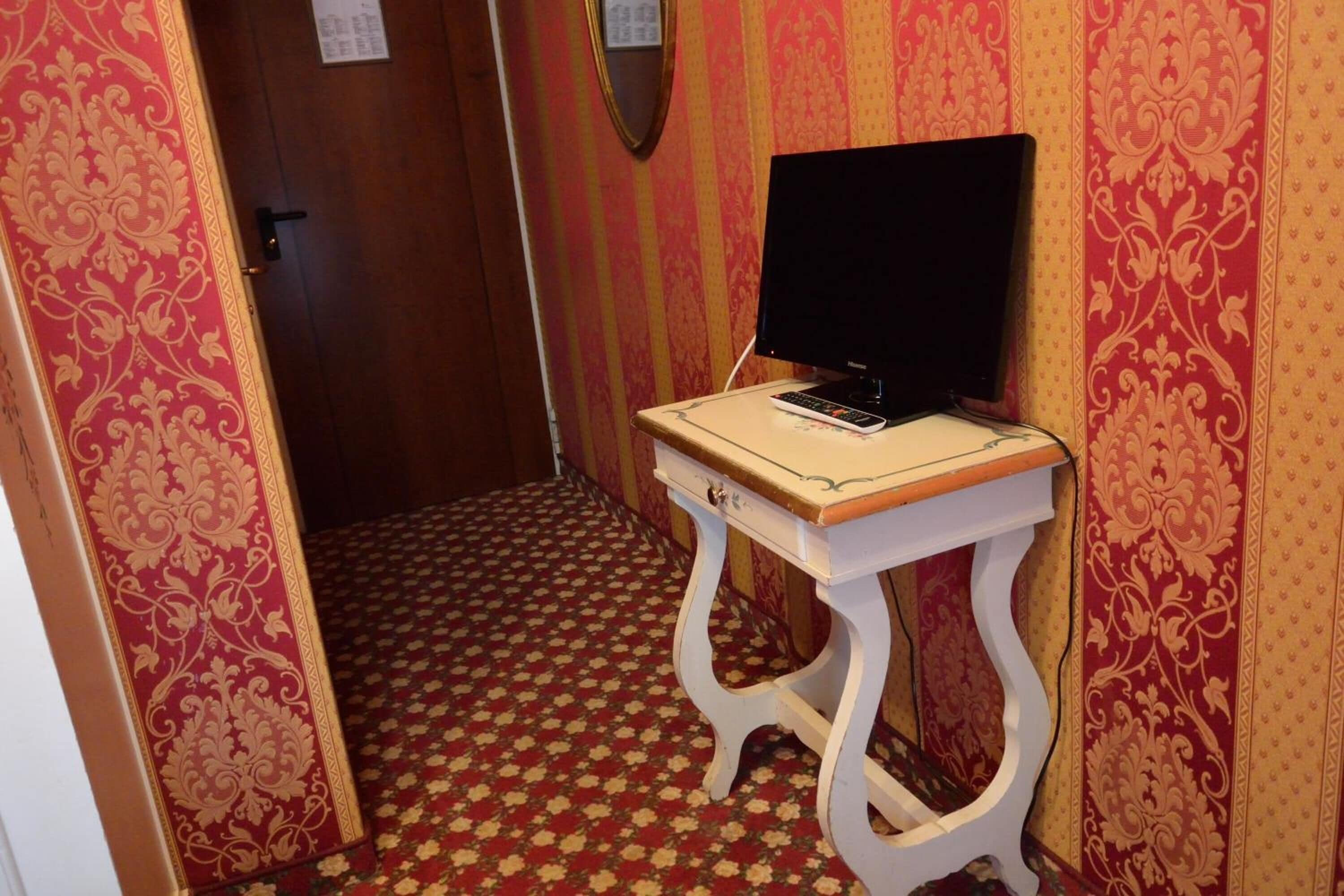 A narrow hallway with patterned wallpaper, a wooden door, and a small table with a computer and remote control.