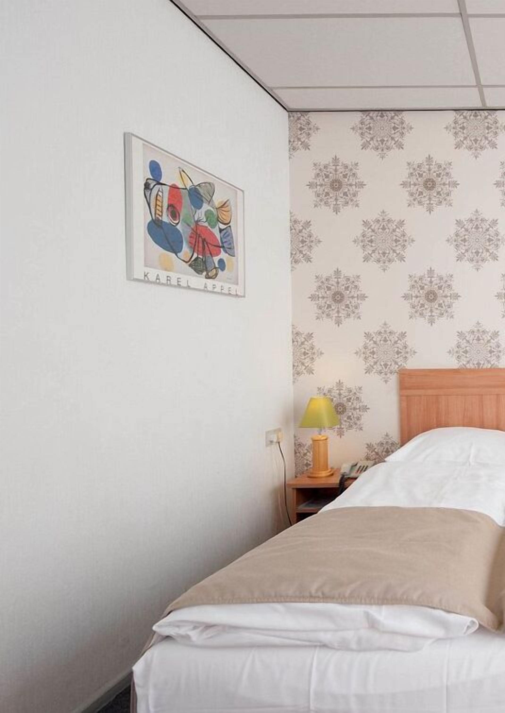 A hotel room with a bed, a nightstand with a lamp, and a wall with a floral wallpaper pattern.