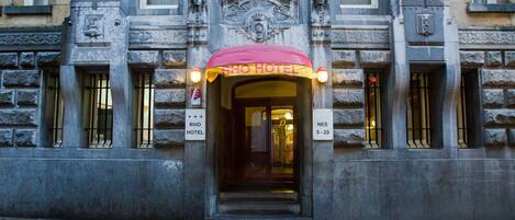 L’entrée de l’hôtel RHOF, reconnaissable à son auvent rouge, sa façade en pierre et un panneau indiquant si des chambres sont disponibles.