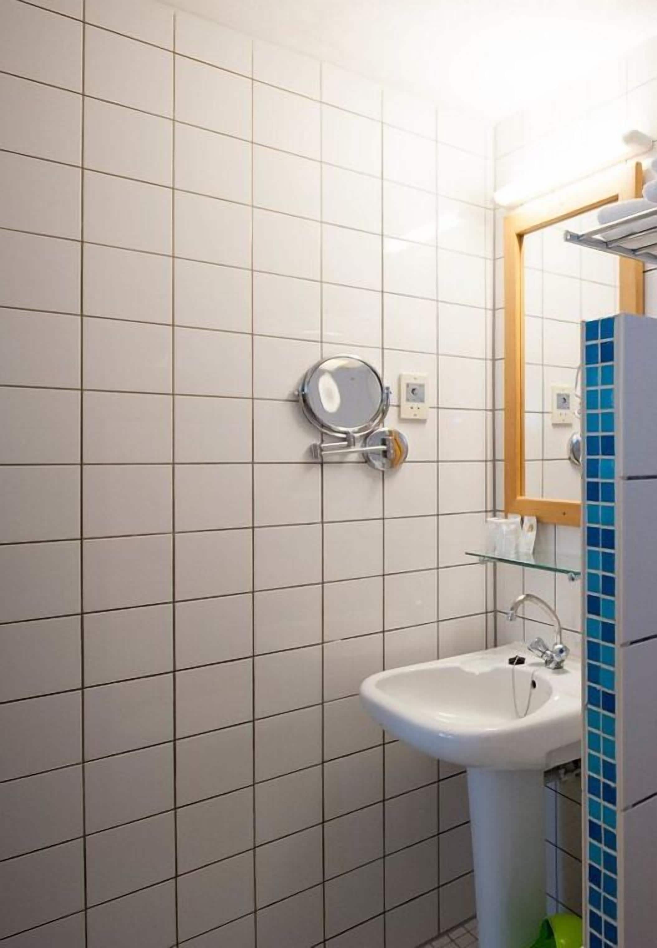 Un baño con paredes de azulejos blancos, un espejo redondo, un lavabo y un estante de vidrio.