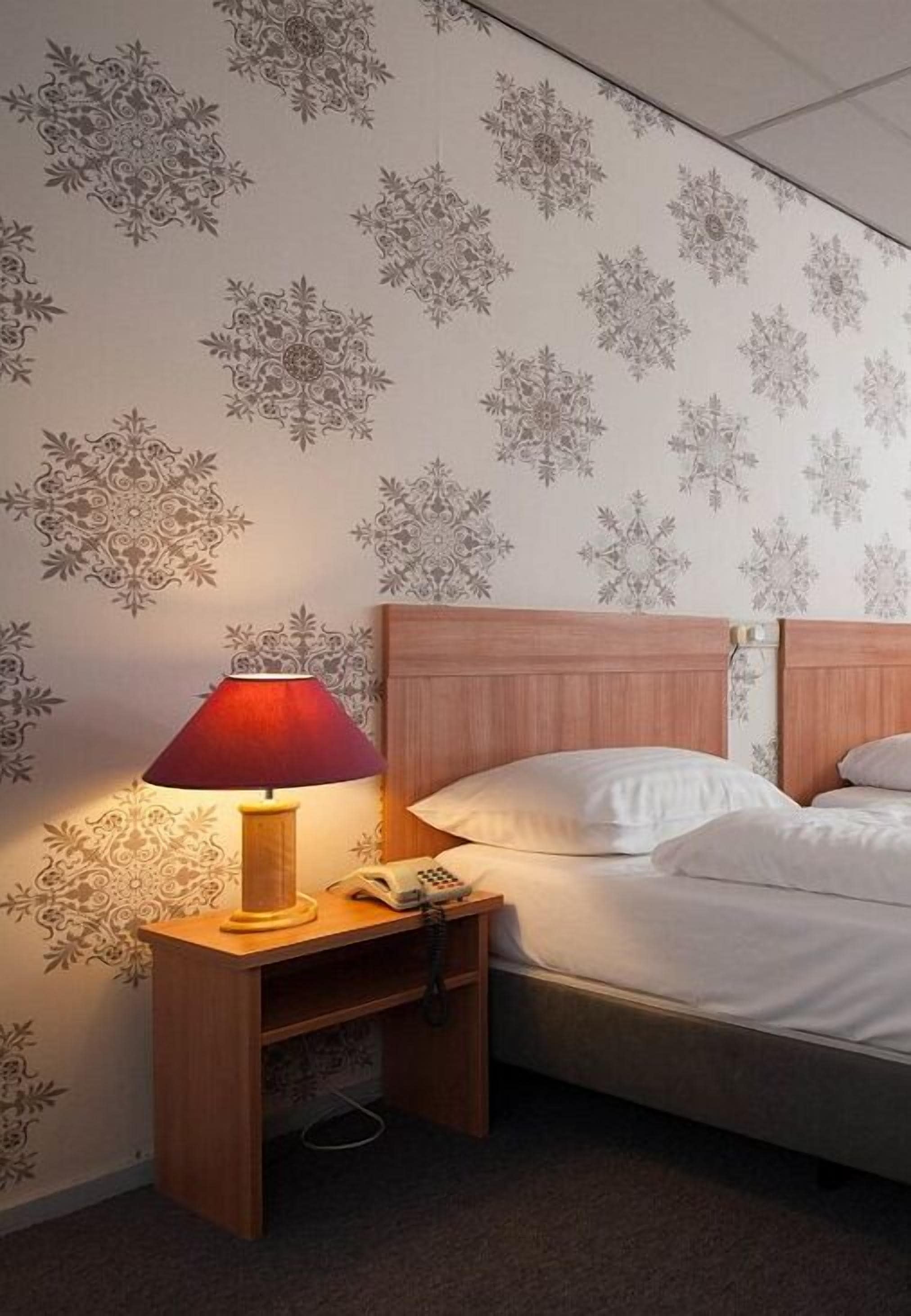 Quarto de hotel com uma cama, uma mesa de cabeceira de madeira com luminária e um telefone.