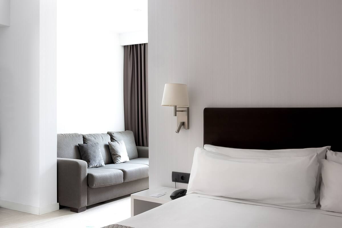 A modern hotel room with a bed, a bedside table, a lamp, a sofa, and a window with curtains.
