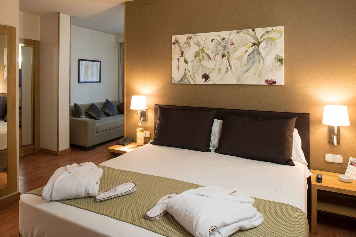 A hotel room with a bed, two bedside lamps, a painting on the wall, a sofa in the background, and a mirror.