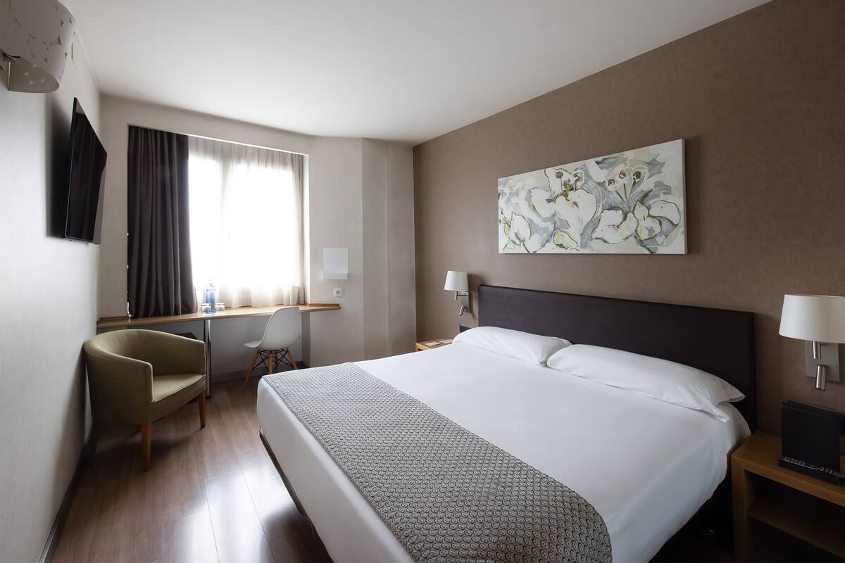 A hotel room with a large bed, a desk with a chair, a TV, and a painting on the wall.