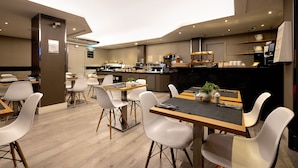 Un comedor de hotel moderno con varias mesas y sillas, una zona de cocina al fondo y un amplio ventanal que proporciona luz natural.