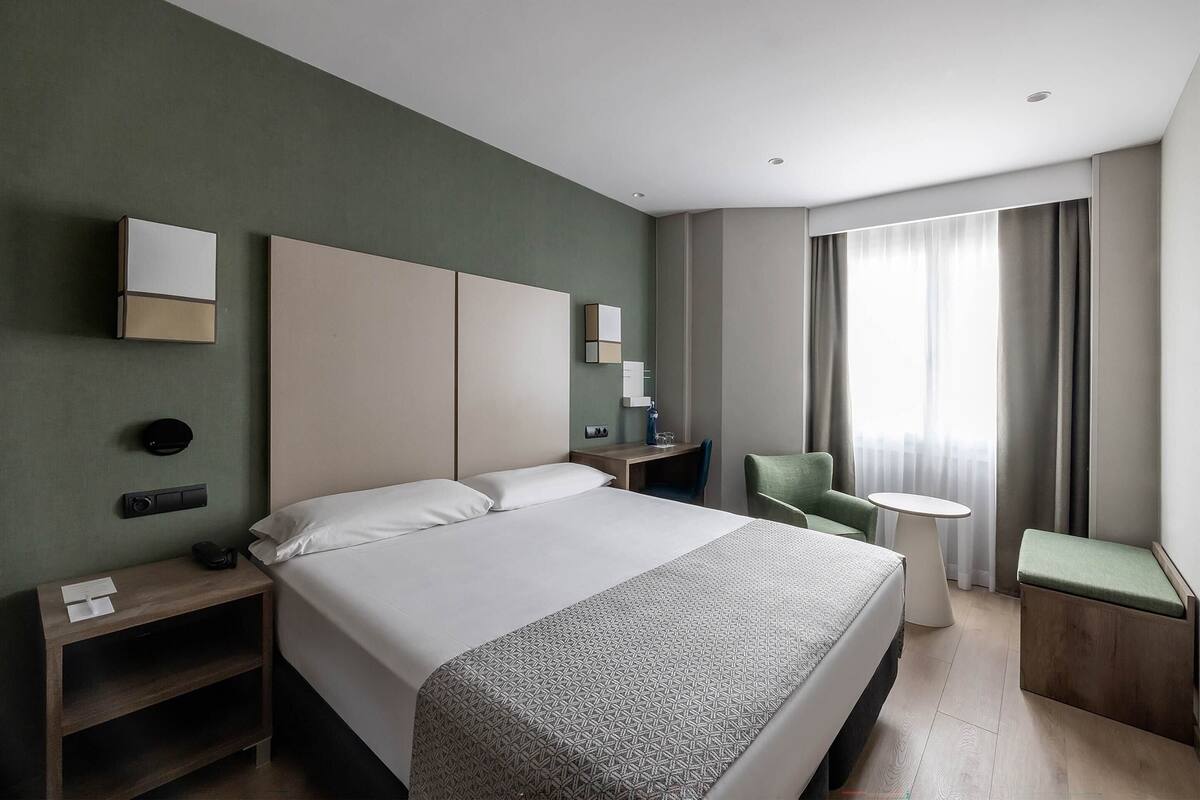 A modern hotel room with a large bed, a desk, a chair, and a small table.