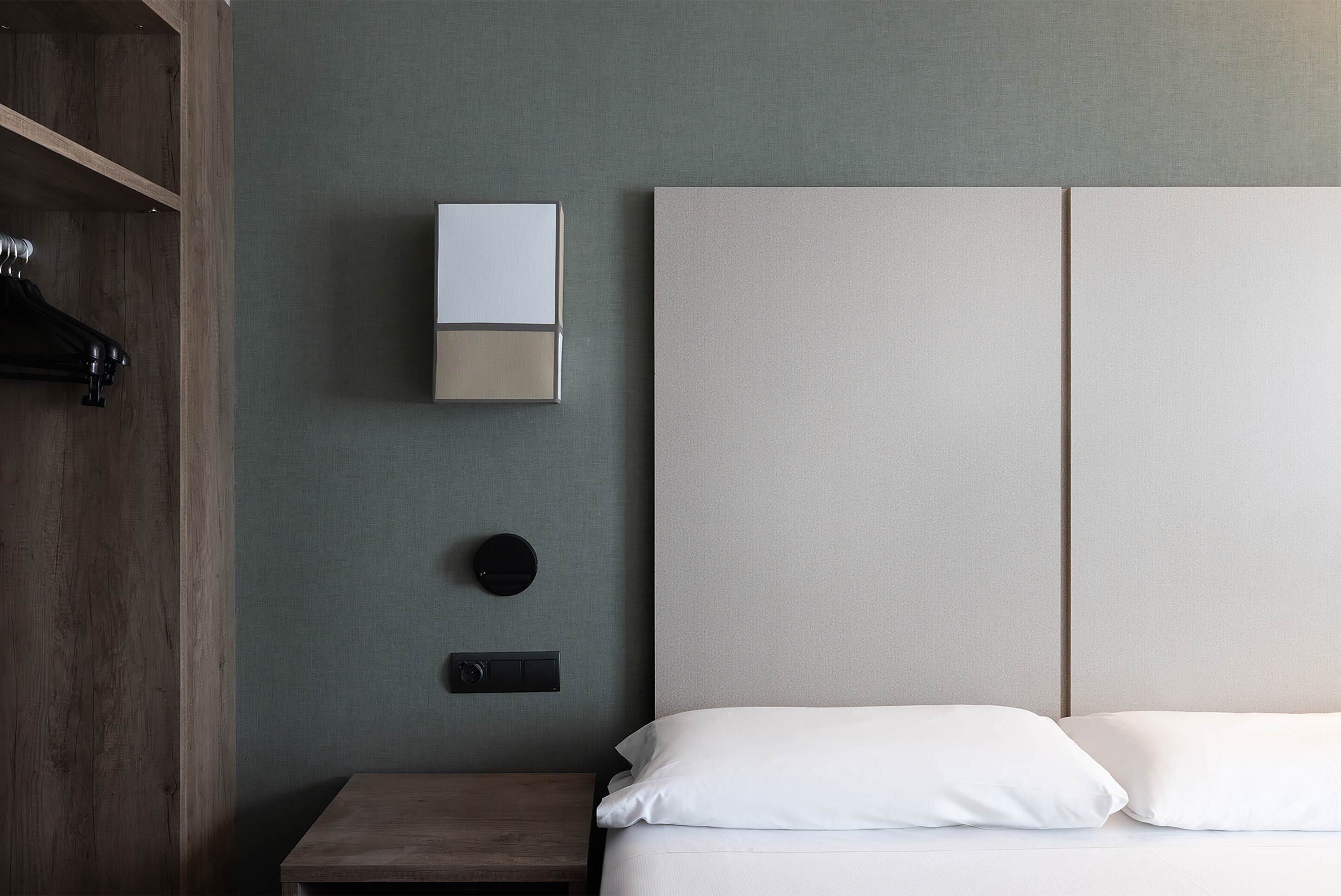 A minimalist hotel room with a bed, a nightstand, a wall-mounted light, and a coat rack.
