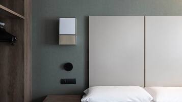 A minimalist hotel room with a bed, a nightstand, a wall-mounted light, and a coat rack.