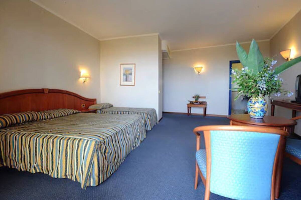 A hotel room with a bed, a chair, a small table, and a vase with flowers.
