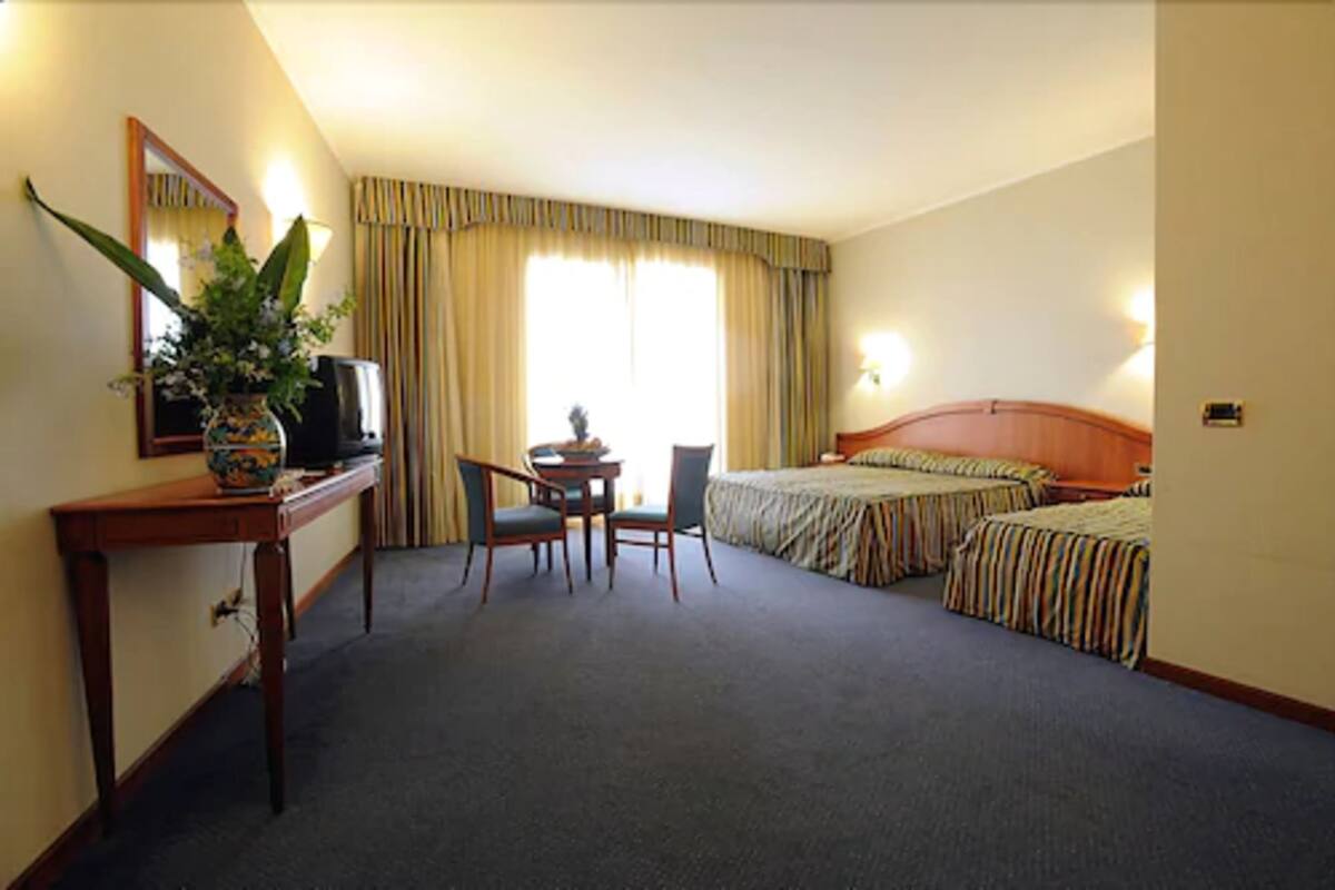 A hotel room with a desk, two single beds, a chair, a TV, and a large window with curtains.