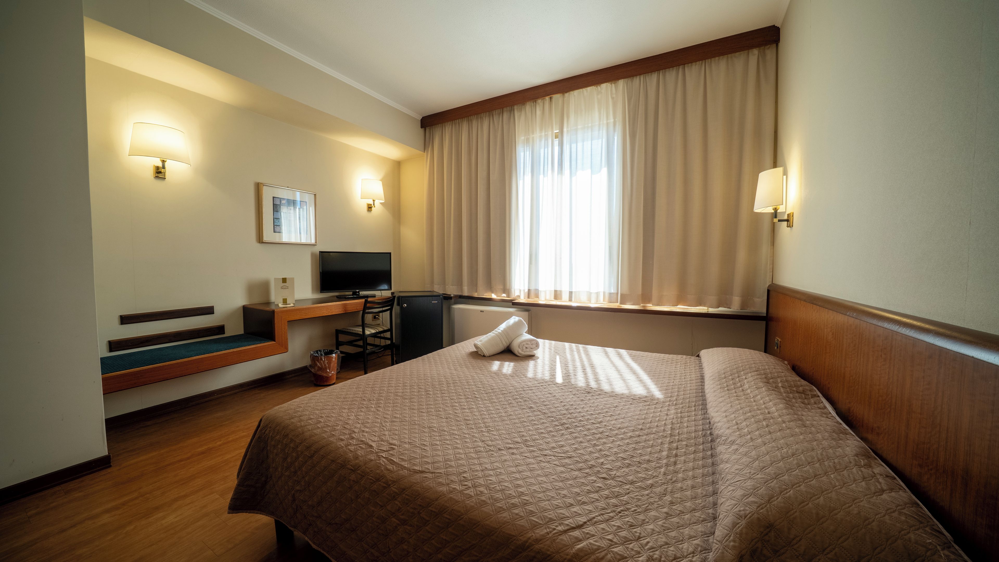 Una habitación de hotel con cama, escritorio, televisor y ventana con cortinas.