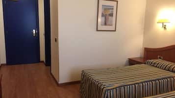 Quarto de hotel com duas camas, uma cabeceira de madeira, uma mesinha de cabeceira, um quadro na parede e uma porta com puxador.