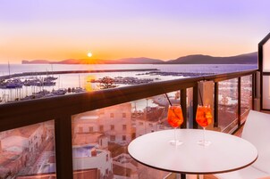 Un balcone con vista su un porto turistico e un tramonto. Sul tavolo ci sono due bicchieri di bevanda rossa.