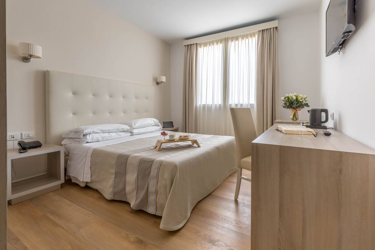 Habitación de hotel con una cama grande, mesitas de noche, un escritorio con silla, televisión y ventana con cortinas.