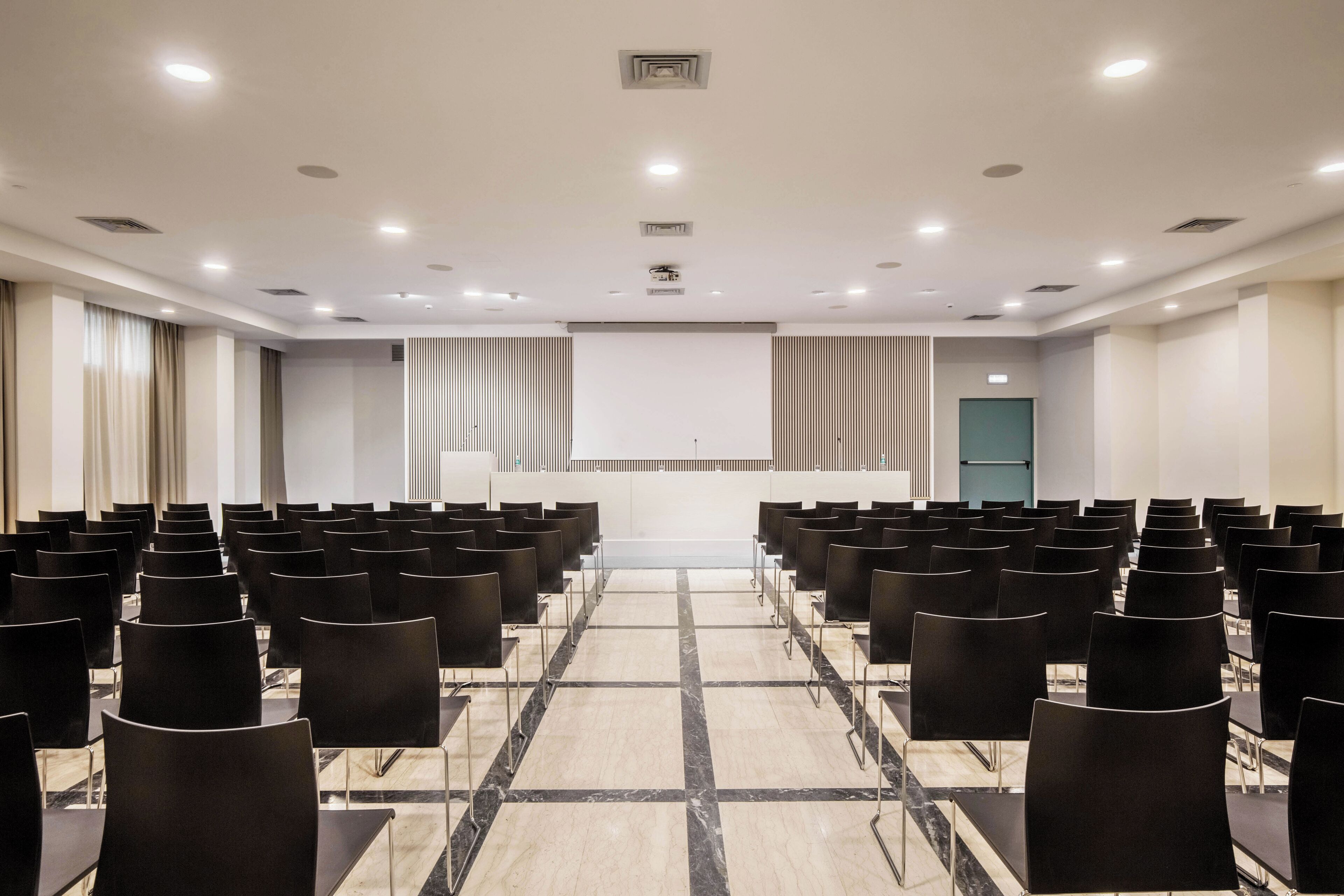Une salle de conférence avec des rangées de chaises noires, une scène et un écran de projection.