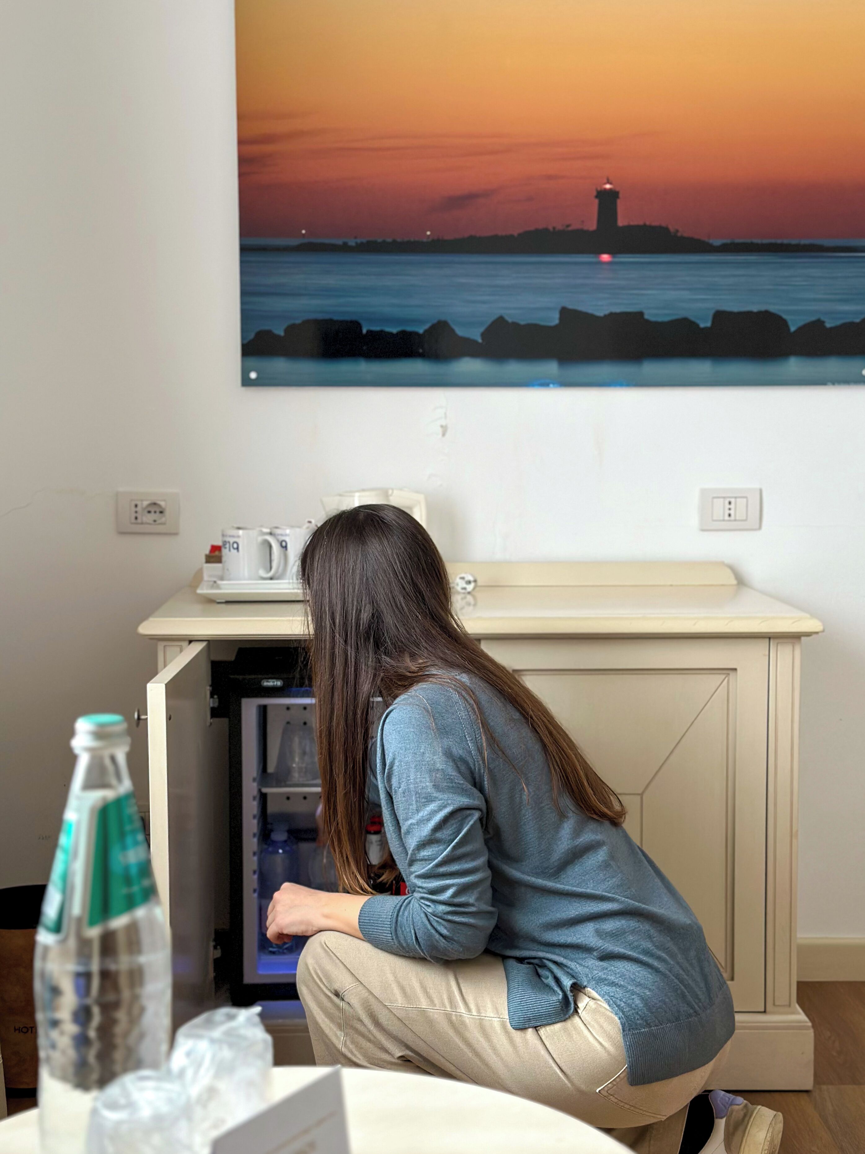 A person in a room with a refrigerator, a bottle of water, and a painting of a sunset over water.