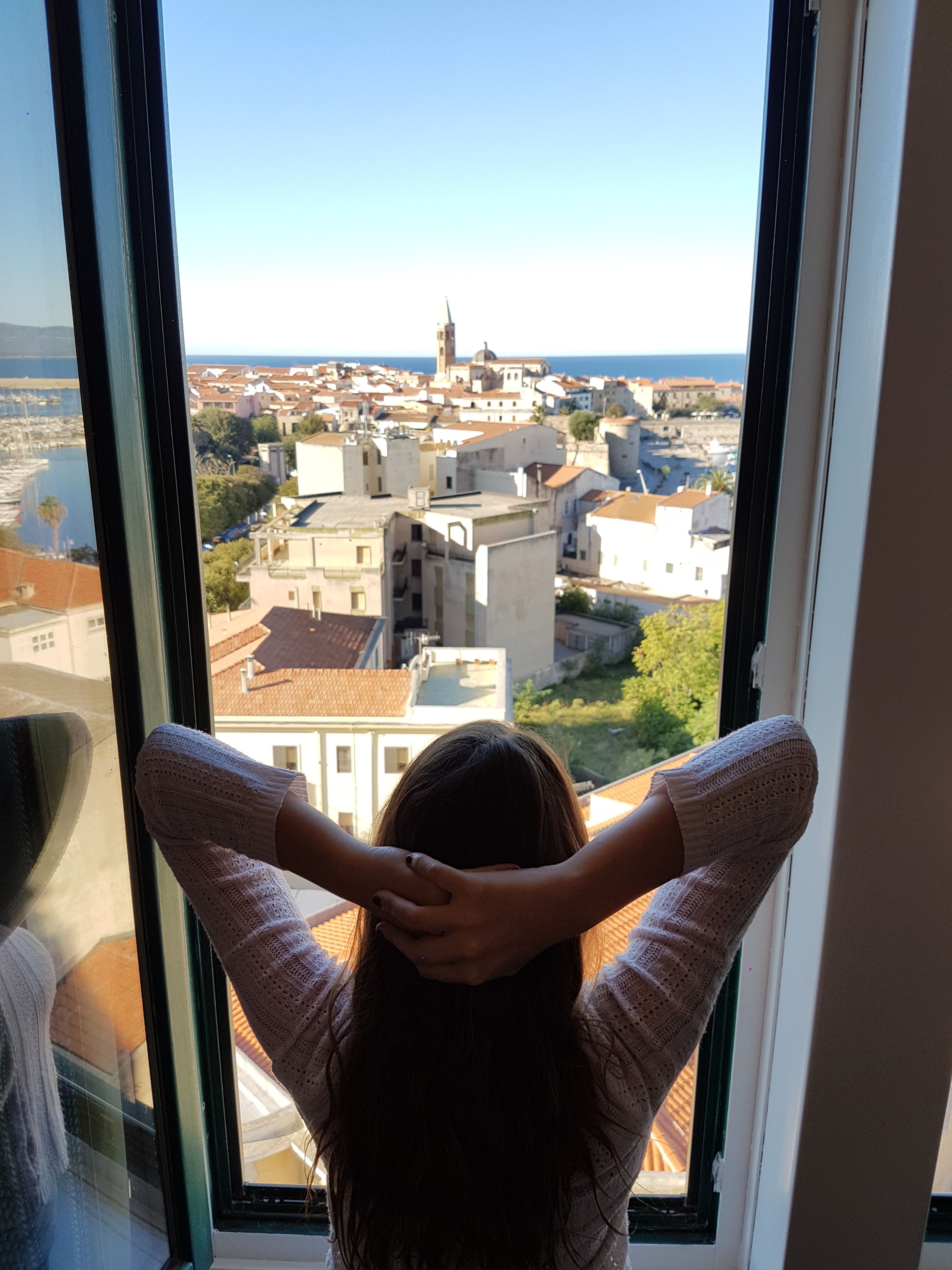A person standing by a window with a view of a coastal town, buildings, and a tower.
