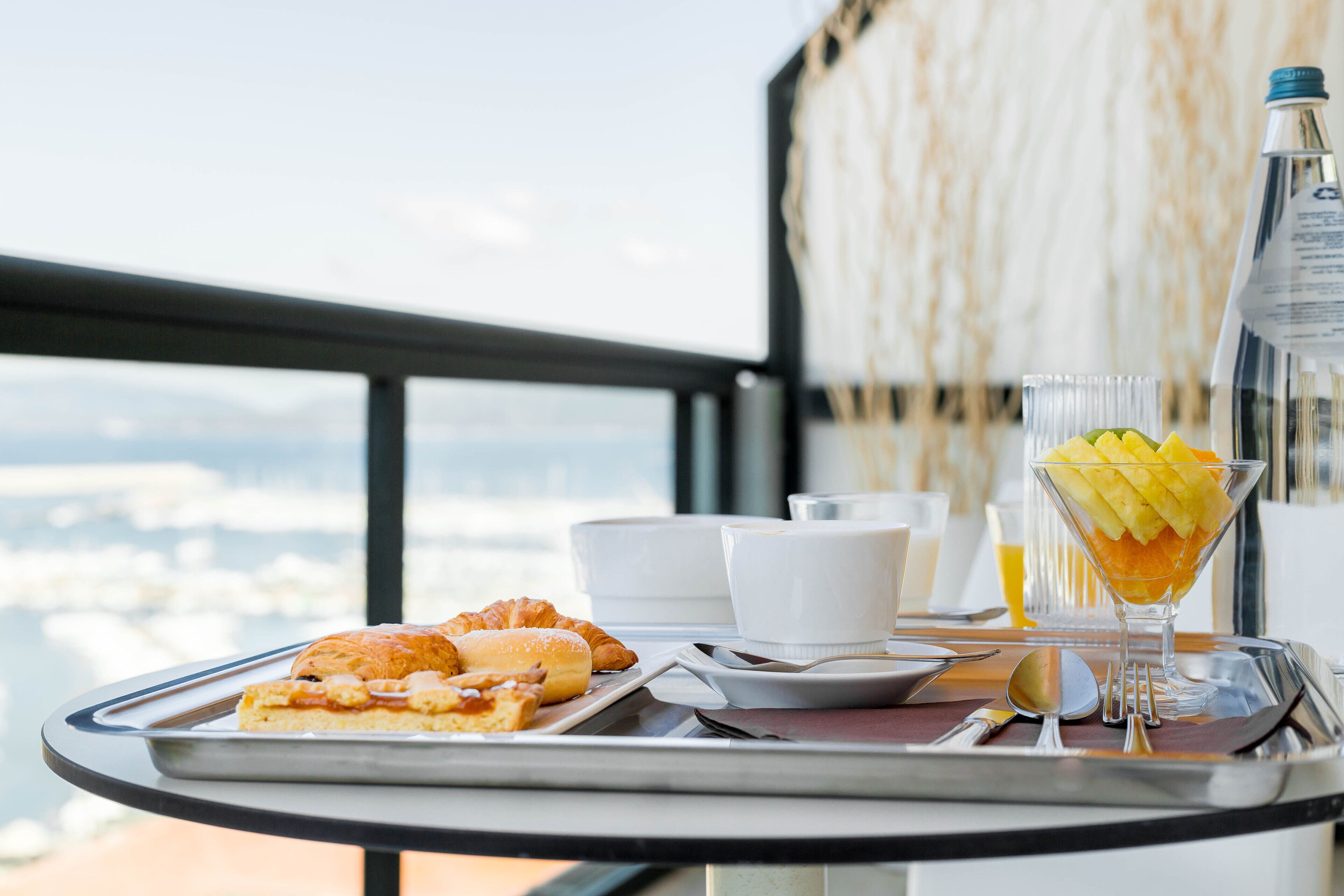 A breakfast tray with croissants, a glass of orange juice, and a fruit bowl on a balcony with a city view.