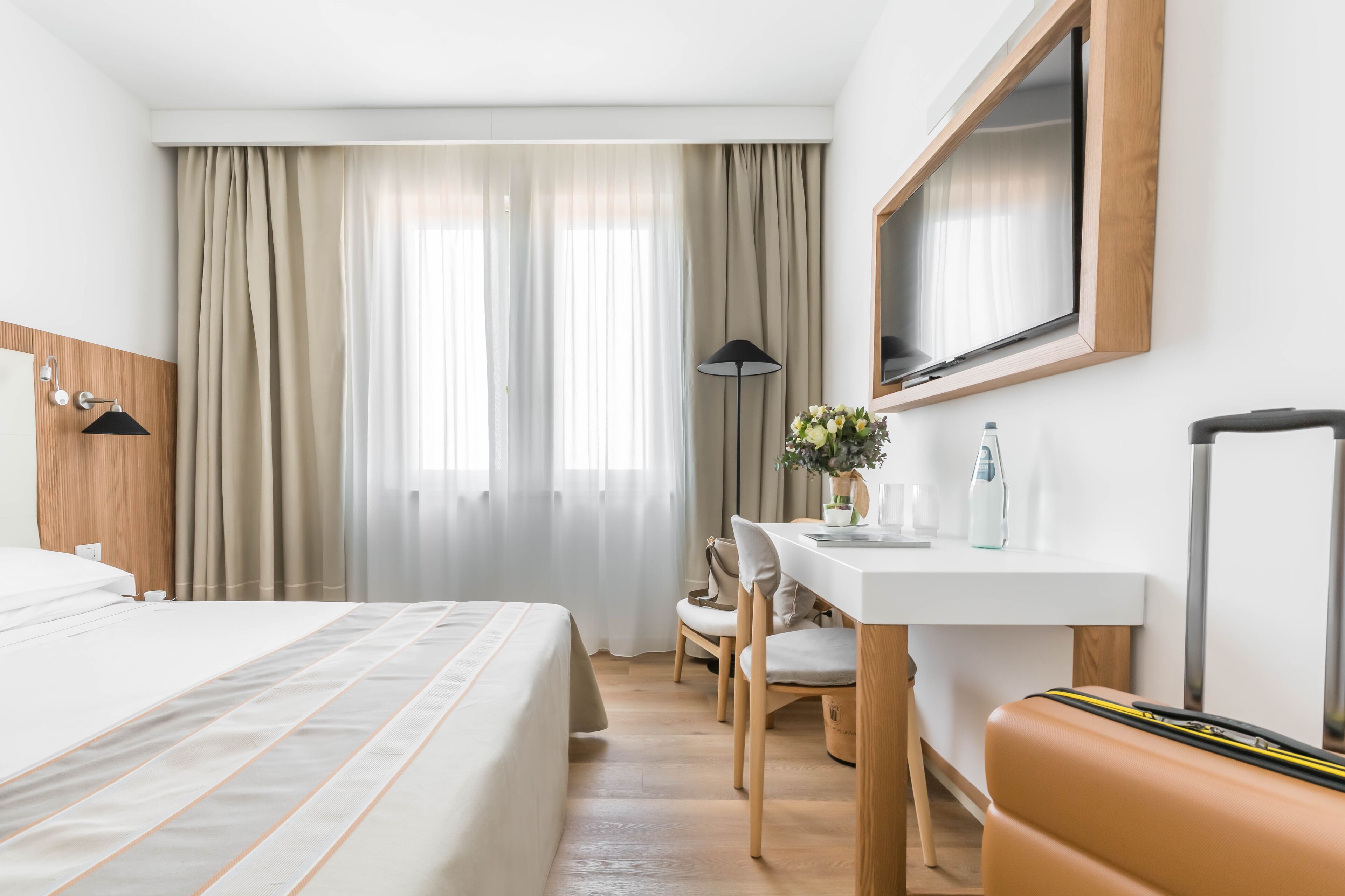 Une chambre d’hôtel avec un lit, un bureau, une chaise, une lampe, une télévision et une valise.