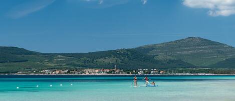A coastal scene with clear turquoise waters, a small beach, and a distant hill with buildings.