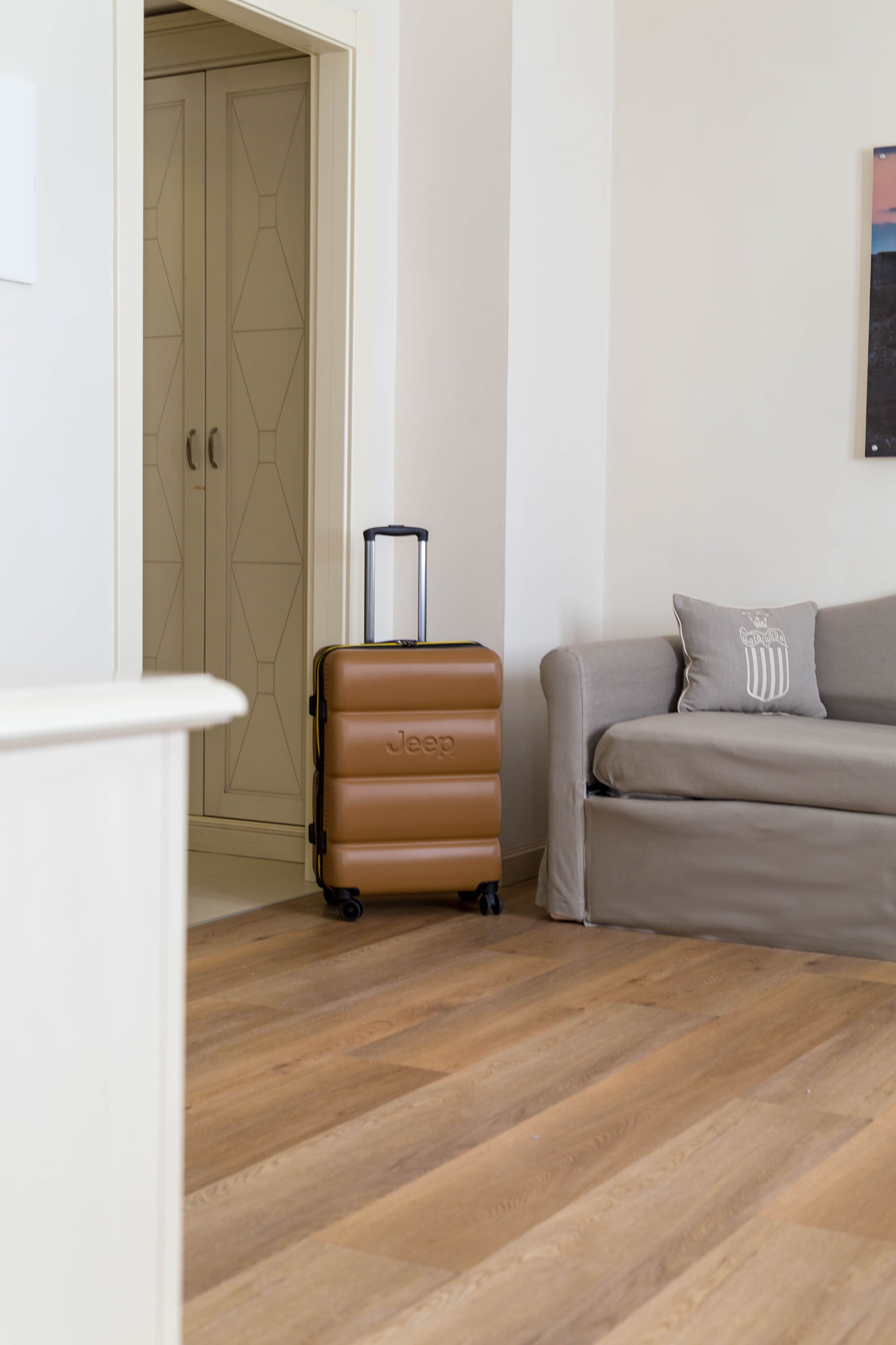 A room with a wooden floor, a brown suitcase, a grey sofa, and a door with geometric patterns.