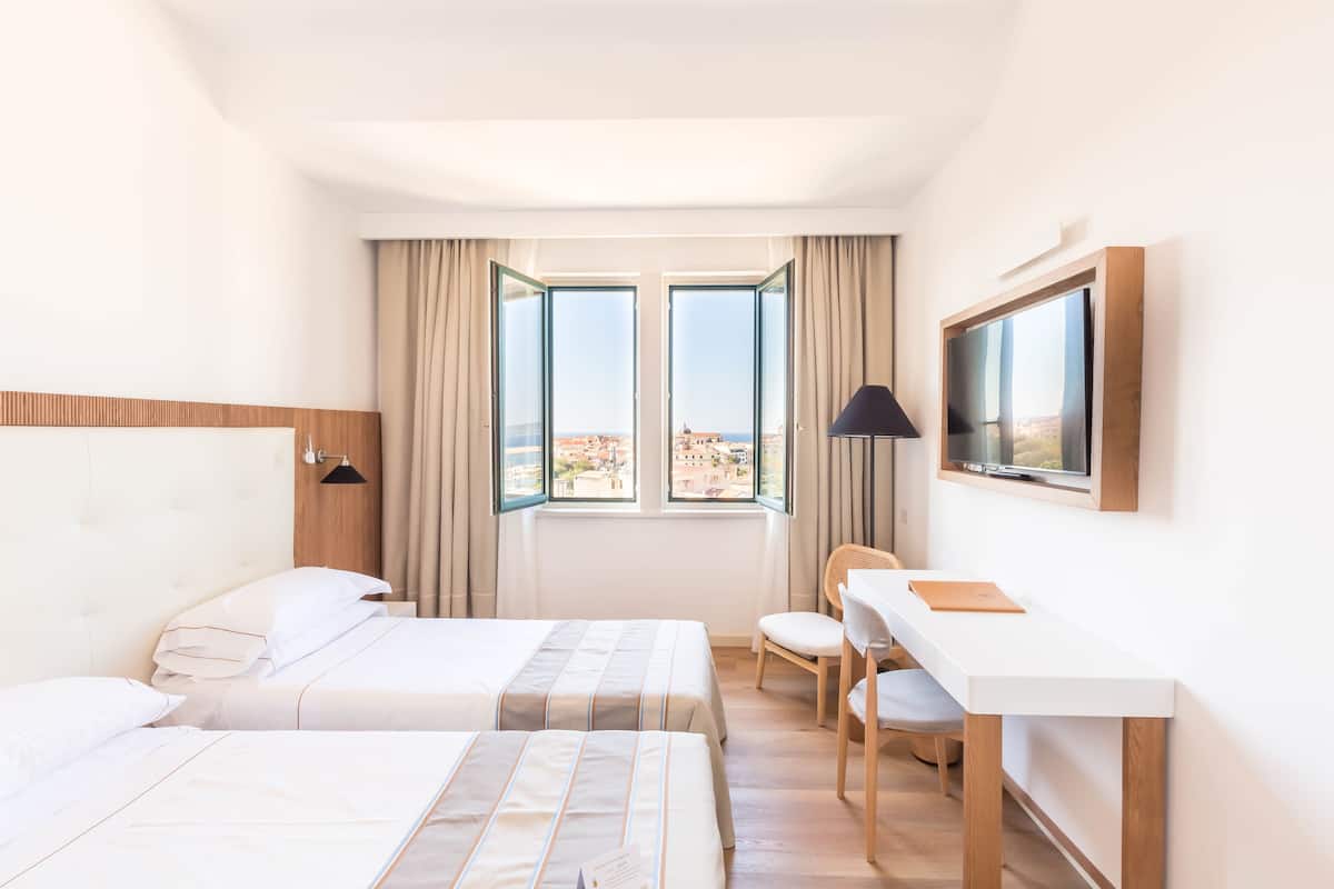 Habitación de hotel con dos camas, un escritorio, una silla, un televisor y ventanas grandes con cortinas.