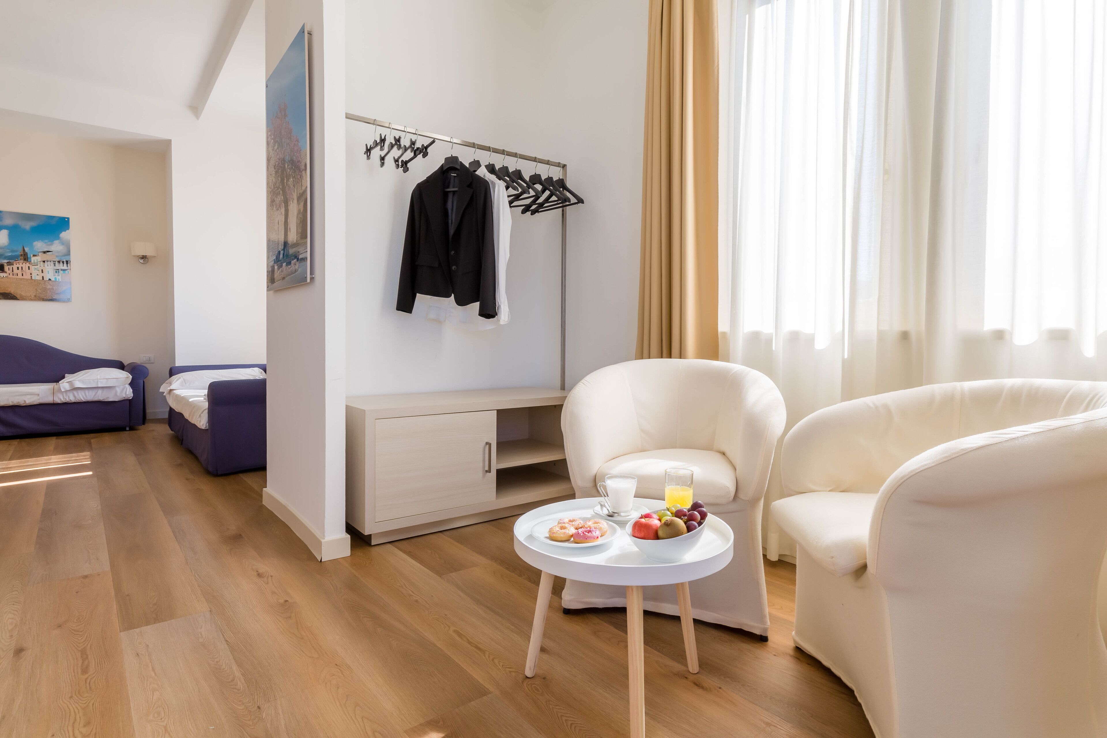 Una camera d'albergo moderna con pavimento in legno, un divano bianco, un tavolo da pranzo con colazione, un appendiabiti e un quadro appeso alla parete.