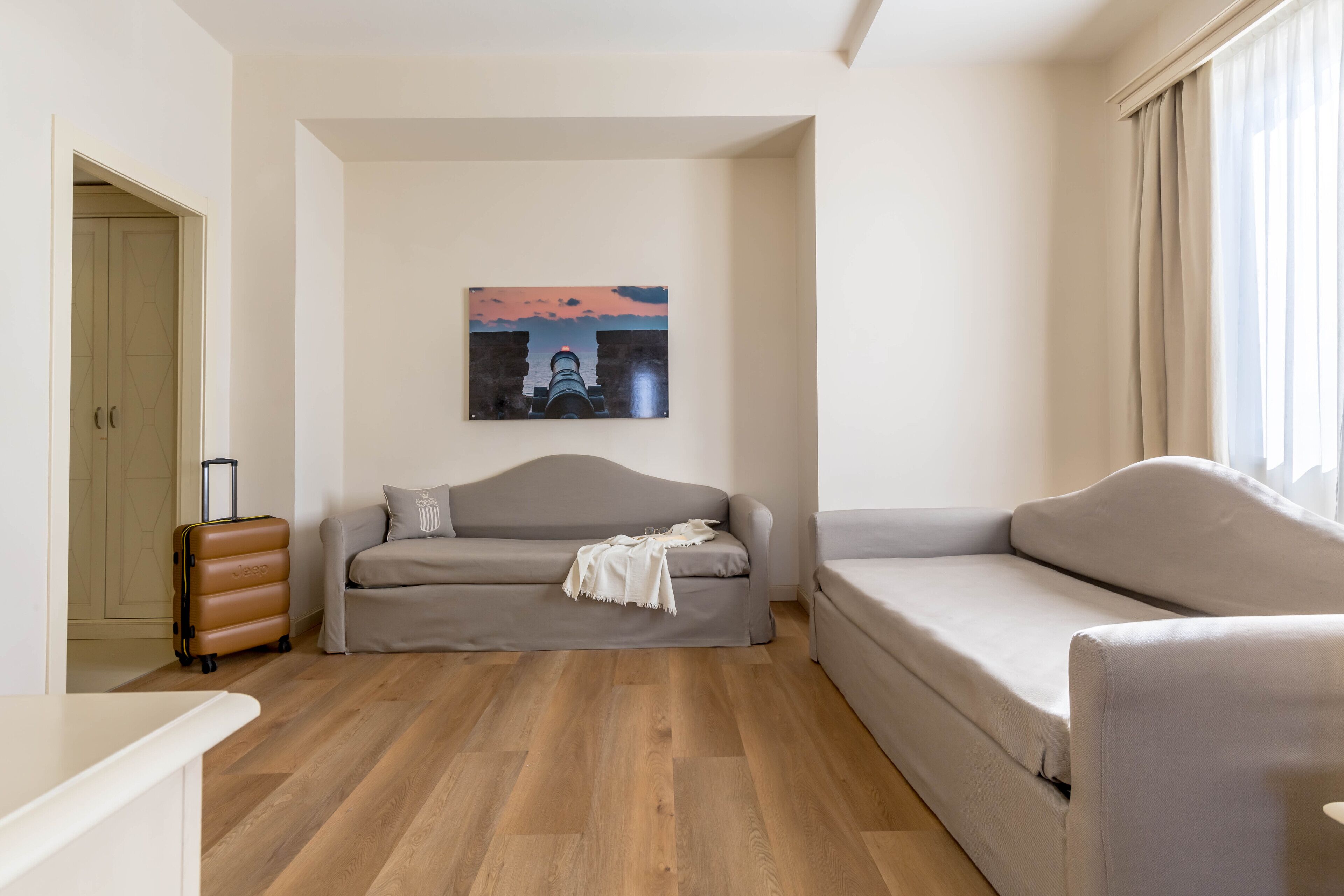 A modern living room with a grey sofa, a smaller grey armchair, a brown suitcase, and a painting of a sunset over a bridge.