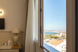 A hotel room with a view of a coastal city, a bedside table with a lamp, and a window with curtains.