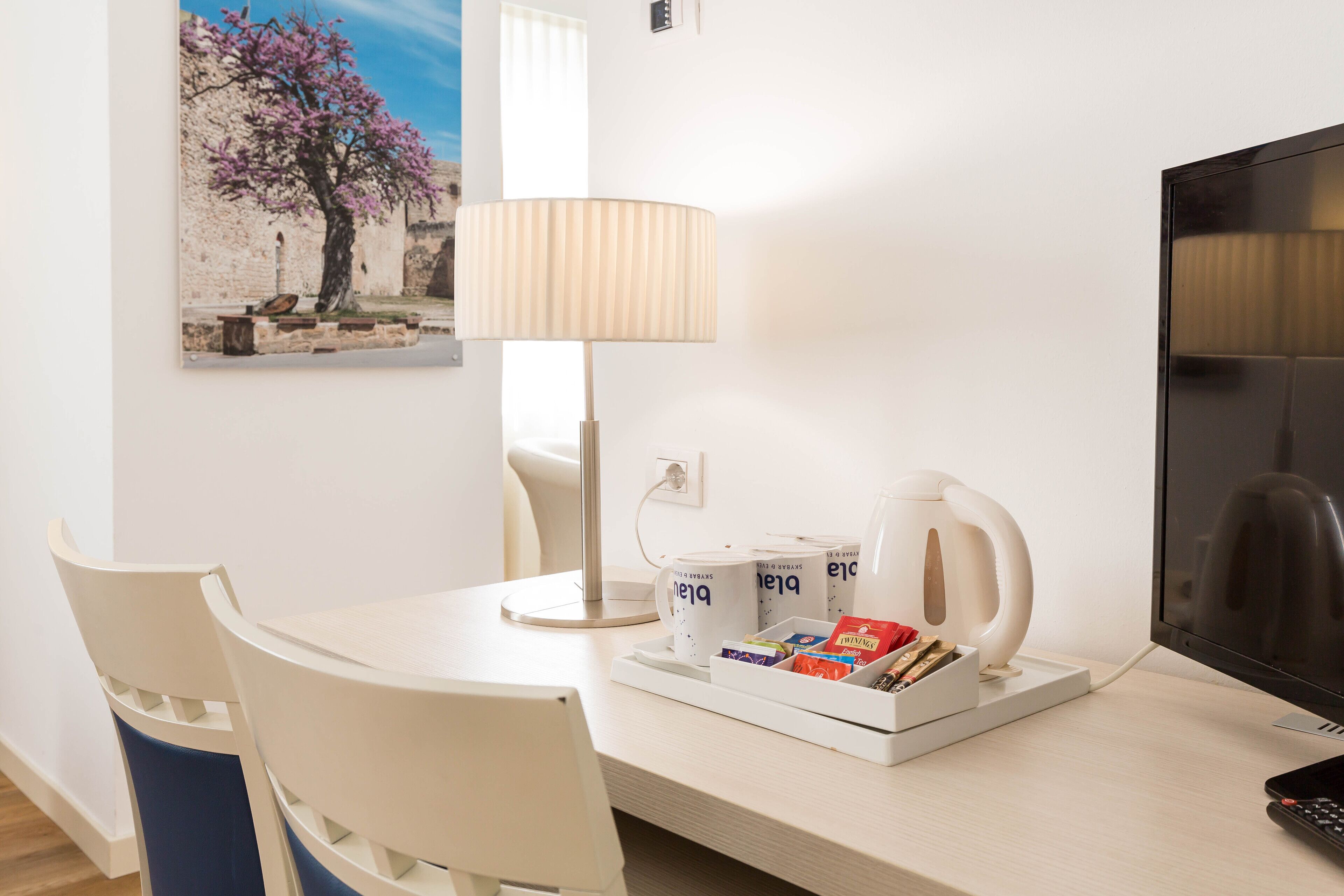 A hotel room with a white desk, a chair, a television, a lamp, and a wall-mounted picture.