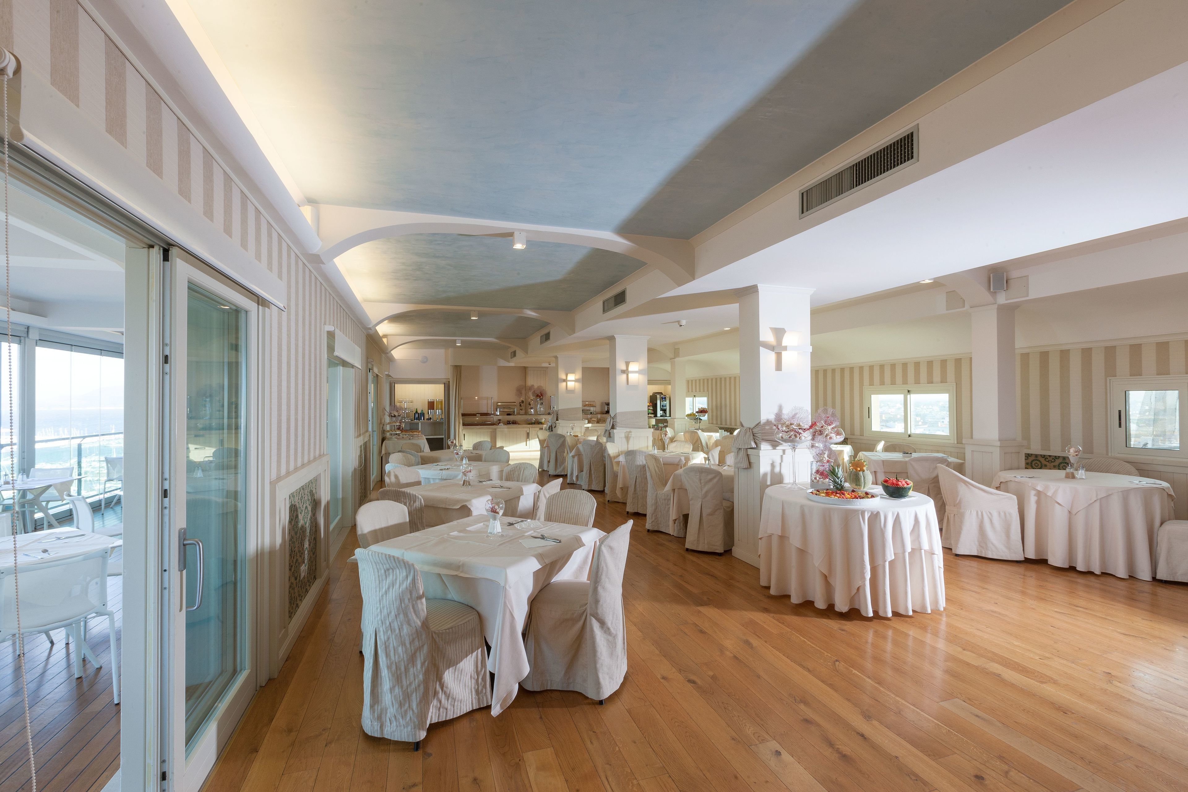 Uma ampla sala de jantar com várias mesas redondas e cadeiras, janelas grandes e um pé-direito alto.