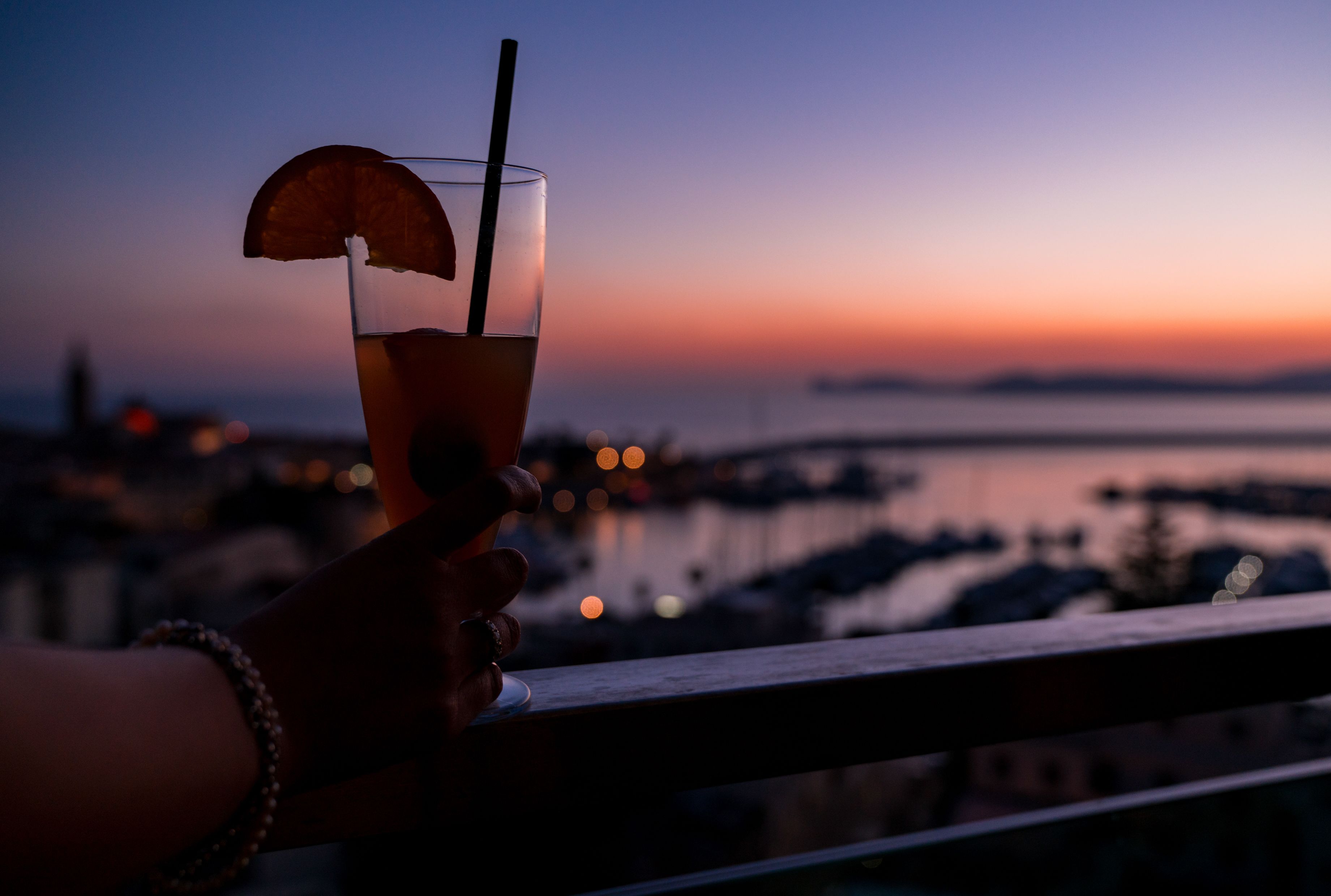 A hand holding a glass with a straw and a slice of fruit, overlooking a marina at sunset.