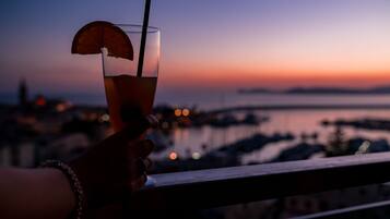 A hand holding a glass with a straw and a slice of fruit, overlooking a marina at sunset.