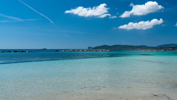 A clear beach with turquoise water, a rocky breakwater, and distant green hills under a blue sky with clouds. - Hotel Catalunya (Alghero)