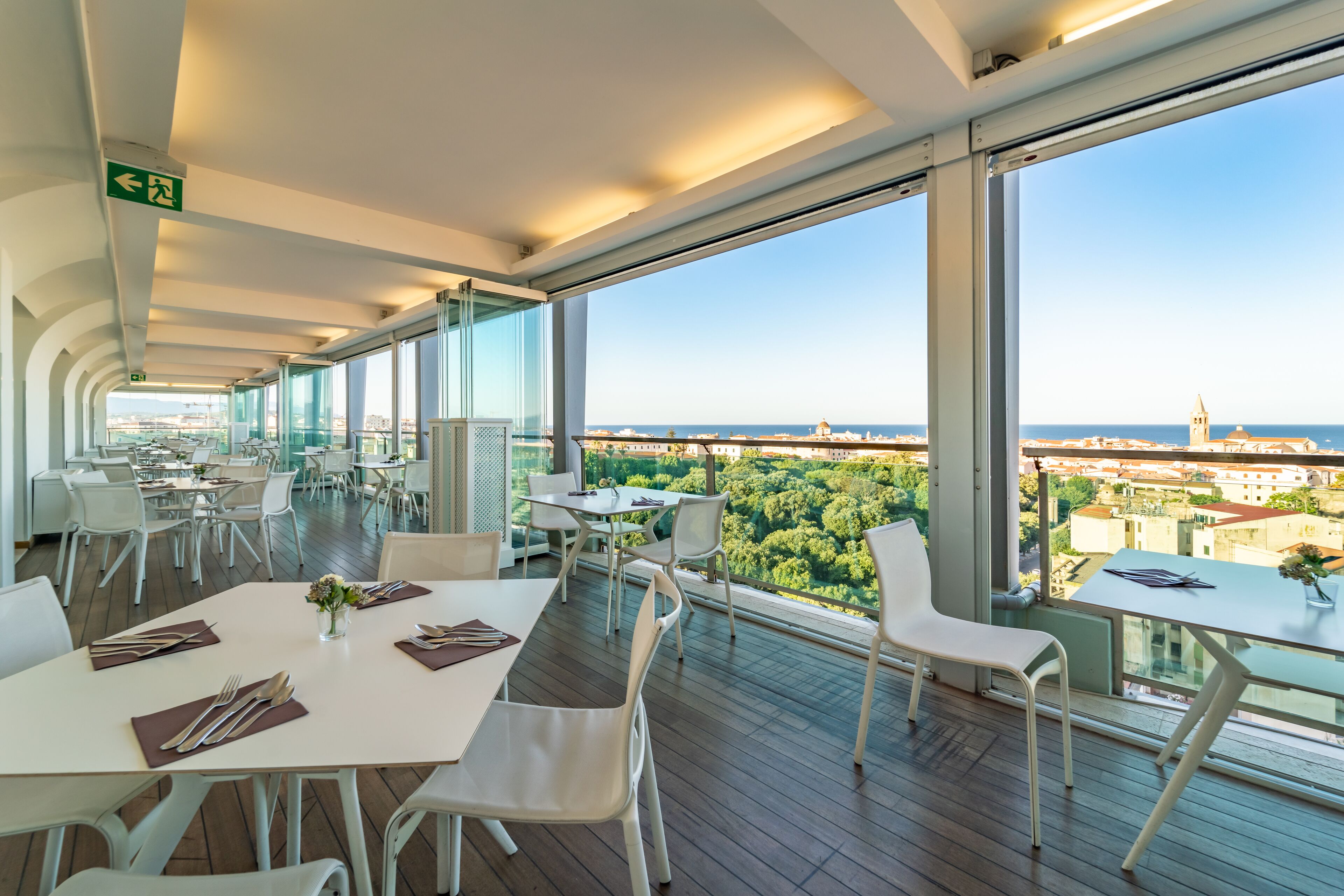 A modern restaurant with white tables and chairs, large windows offering a city view, and a clear sky.