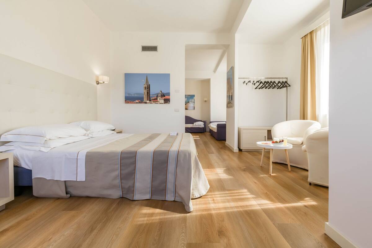 Una habitación de hotel moderna con una cama grande, una zona de estar con una silla y una mesa, y vistas a un paisaje urbano a través de la ventana.